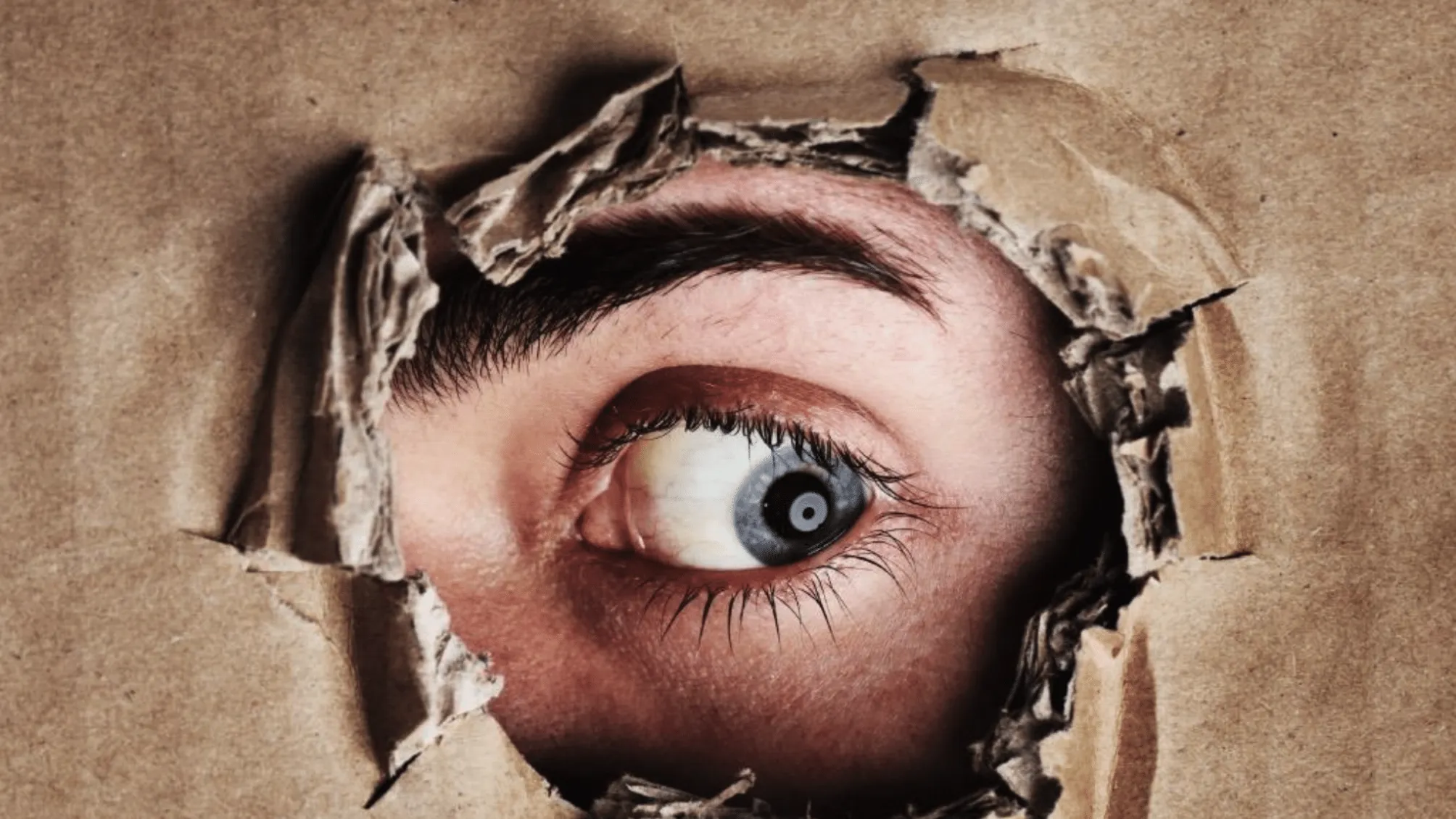 A close-up of a human eye peering anxiously through a jagged hole torn in brown cardboard.